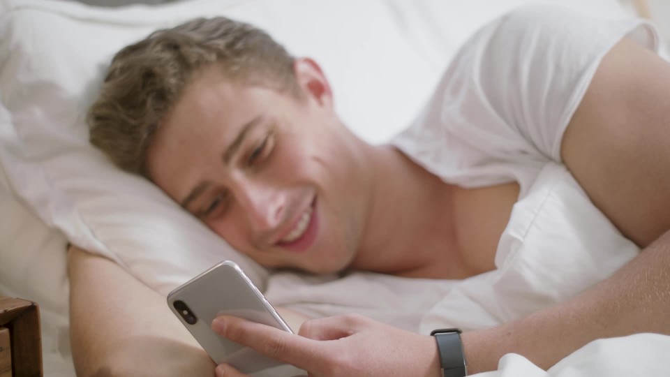250ae88b 1c61 4139 8311 6d1d8157db90.thumbnail Handsome man sending funny text message on cell phone to friend relaxing in bed | ClipStock
