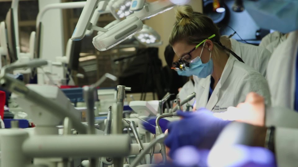 Female and male workers sculpting dental prosthetics at workbench under lights from behind